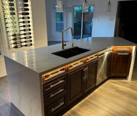 Elegant kitchen island featuring a Calacatta quartz waterfall countertop, dark wood cabinetry with underglow lighting, a gold faucet, black double sink, and an LED-lit wall-mounted wine rack, complemented by crystal pendant lights.