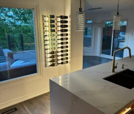 Contemporary kitchen featuring a Calacatta quartz waterfall island, underglow cabinet lighting, gold faucet, crystal pendant lights, and an LED-lit wall-mounted wine rack with a scenic outdoor view.