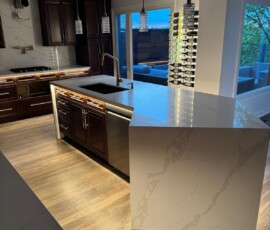A contemporary kitchen with a large island featuring luxurious Calacatta quartz countertops, dark wood cabinets, underglow lights beneath the island, stainless steel appliances, pendant lighting, and hardwood flooring, complemented by a wall-mounted wine rack near the sliding glass doors.