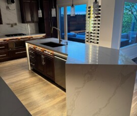 A contemporary kitchen with a large island featuring luxurious Calacatta quartz countertops, dark wood cabinets, underglow lights beneath the island, stainless steel appliances, pendant lighting, and hardwood flooring, complemented by a wall-mounted wine rack near the sliding glass doors.