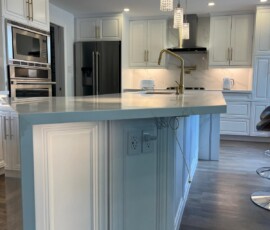 Elegant white kitchen island featuring a quartz waterfall edge, mitered corners, built-in electrical outlets, gold faucet, and crystal pendant lighting.