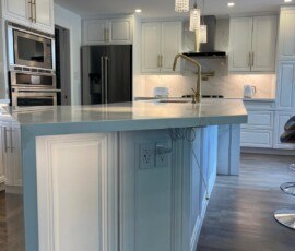 Elegant white kitchen island featuring a quartz waterfall edge, mitered corners, built-in electrical outlets, gold faucet, and crystal pendant lighting.