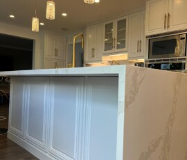 Close-up of a luxurious kitchen island featuring a quartz waterfall leg with mitered edges, custom paneling, and gold fixtures, illuminated by modern crystal pendant lights.