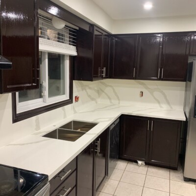 A sleek kitchen with dark glossy cabinets, white quartz countertops, a stainless steel sink, stainless steel appliances, and tiled flooring, illuminated by recessed ceiling lights.