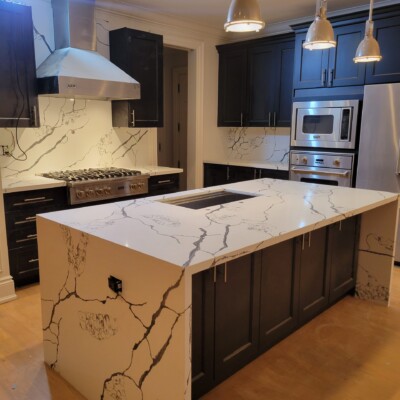 A modern kitchen with a large island and striking Calacatta quartz countertops featuring bold veining, complemented by dark cabinetry, stainless steel appliances, a range hood, and pendant lighting, with matching quartz backsplash for a seamless, elegant look.