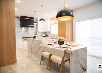 Open-concept kitchen and dining space featuring Calacatta Aurora Gold Quartz waterfall island and matching dining table with bold gold veining, paired with white cabinetry, wood accents, and modern black-and-gold lighting.