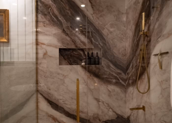 Interior of a luxury shower featuring book matched Lyra quartz walls, recessed niche, gold fixtures, and a linear drain.