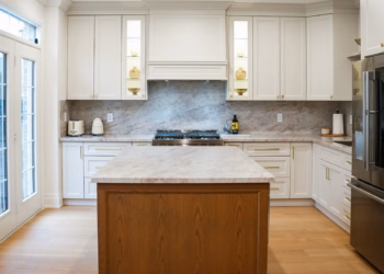Taj Mahal Quartz full-height backsplash with warm veining behind a kitchen range, paired with white cabinets and brass hardware.