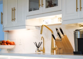 Calacatta Oro quartz backsplash shown behind a gold kitchen faucet, with white cabinetry and under-cabinet lighting.